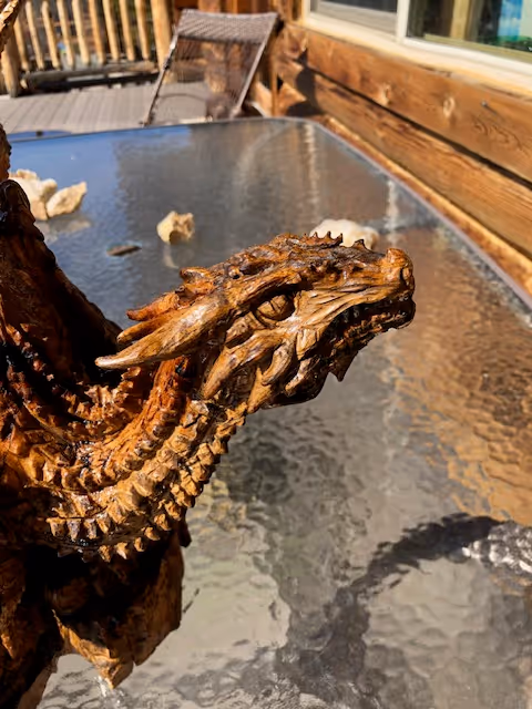 A close-up of one of the dragon heads from a wooden sculpture. The carving features sharp details, with fierce eyes, layered scales, and textured wood grain.